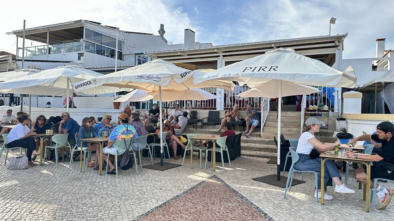 Esplanada do restaurante com chapéus de sol brancos, mesas cheias e a casa de praia ao fundo